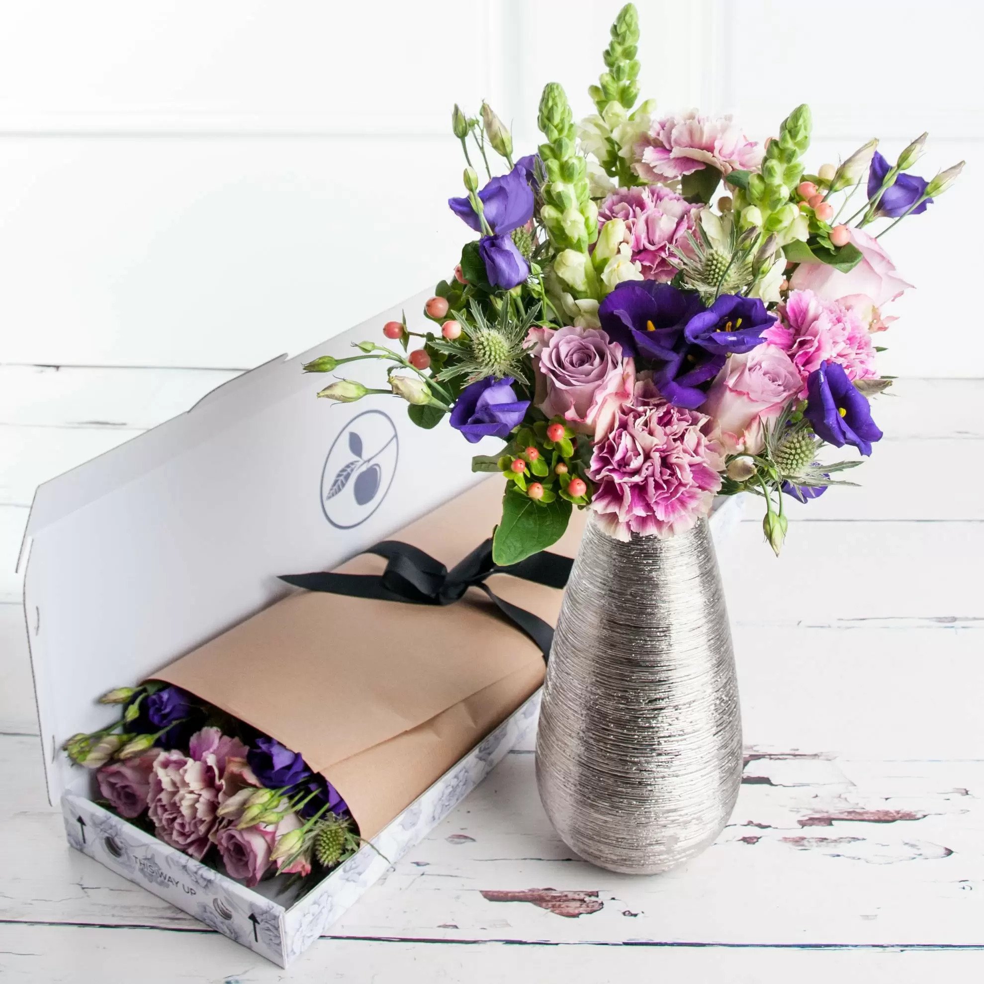 A letterbox bouquet of pink carnations, purple lisianthus, snapdragons, and lilac roses, displayed in a vase next to letterbox packaging.